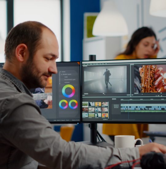 Video maker editing movie using post production software working in creative start up agency office with two monitors. Videographer processing audio film montage on professional computer, taking notes